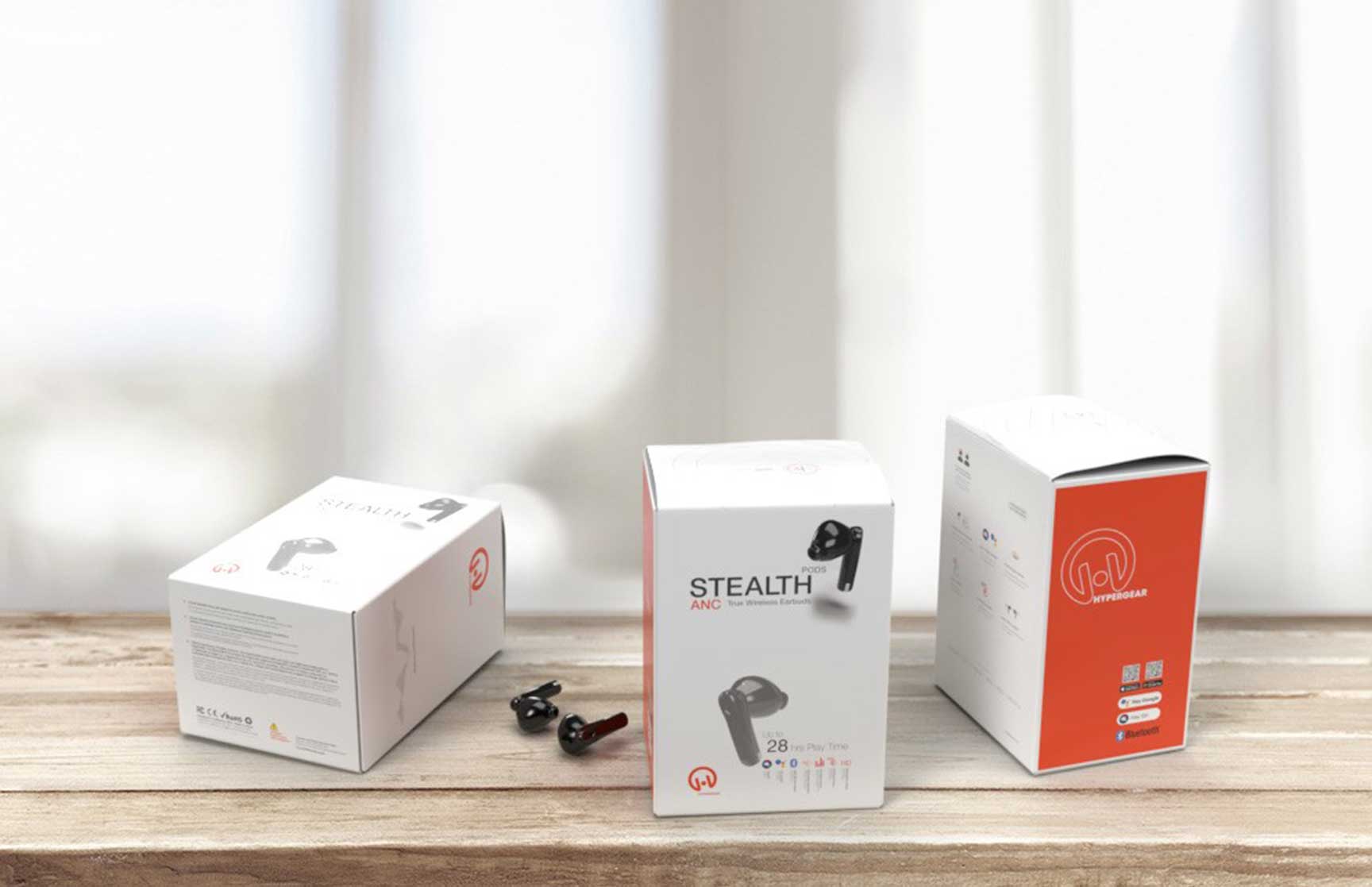 Photorealistic mockup of HyperGear Stealth Pods ANC packaging displayed on a wooden table. Three product boxes are arranged in a staggered layout with the central box showcasing the front panel, including product branding, key features, and a sleek product image. The left box highlights the side panel with product specifications, while the right box displays the back panel featuring the HyperGear logo and additional product information. Two black earbuds are placed in front of the central box. The background features a softly blurred window with natural light creating a clean, minimalistic aesthetic.