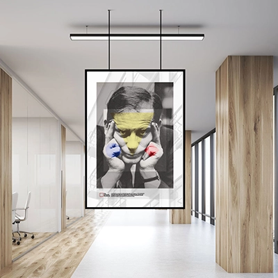 A modern poster design displayed in a minimalistic office setting. The poster features a grayscale portrait of a man holding his head with both hands. His face is accented with bold geometric shapes, including a yellow overlay on his forehead, blue highlights on his right hand, and red highlights on his left hand. The background includes subtle architectural lines, emphasizing clean and structured aesthetics.