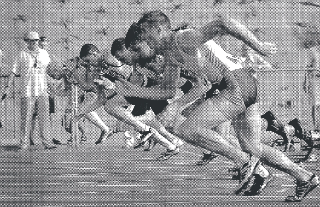 A grayscale photograph of athletes in the middle of a race start, showcasing intense motion and focus. The image captures the runners in a crouched sprinting position, with a background featuring spectators and a track field, emphasizing dynamic movement and athletic determination.
