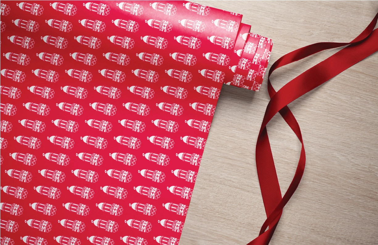 A roll of vibrant wrapping paper featuring a repeated pattern of the Zurich 2038 Winter Olympic Games logo in white against a bold red background. The logo includes a stylized dome structure, the text ‘Zurich 38,’ and the iconic Olympic rings. A red satin ribbon is artistically placed alongside, adding a festive and elegant touch to the composition.